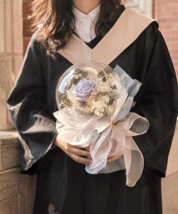 女畢業生穿著黑色學士袍，雙手捧著藍紫玫瑰透明氣球永生花束，呈現溫柔奶油色校園畢業花禮情境。