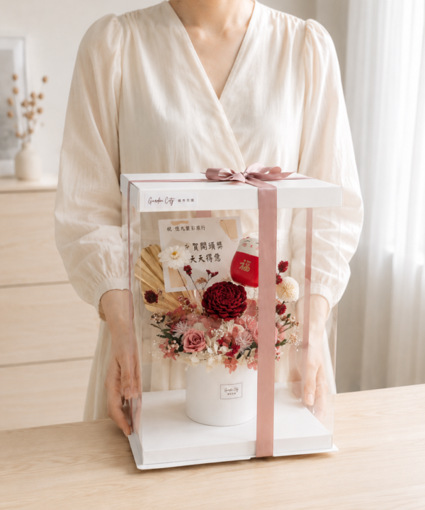 台灣女生雙手展示招財貓永生花盆花禮盒，透明盒搭配粉色緞帶、紅色與粉色永生花材，奶油色自然光電商情境照