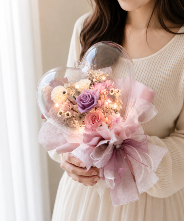 台灣女生雙手捧著粉色愛心波波球永生花束，搭配永生玫瑰、乾燥花與暖色燈串，適合情人節告白與生日送禮