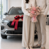 夫妻手拿粉色賀汽車交車花束站在新車旁，灰色汽車搭配紅色交車緞帶，背景為明亮簡約的汽車展示中心。